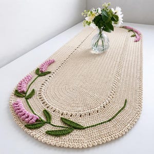 May include: An oval-shaped, cream-colored crochet table runner featuring textured, raised lavender sprigs in light pink with green leaves stitched along the curved edges. The runner has a scalloped border and a dense, uniform stitch pattern.