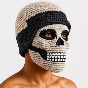 May include: A beige and black crocheted skull mask hat featuring a detailed design with black eye sockets, a black nose cavity, and white teeth. The headgear covers the top of the head and the face, constructed with a textured yarn stitch pattern.