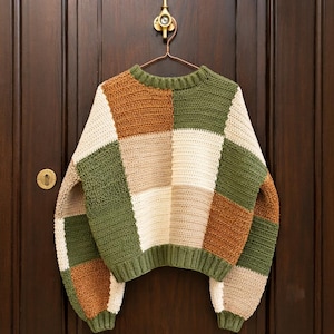 May include: A long-sleeve crochet sweater featuring a patchwork design of square blocks in olive green, cream, and burnt orange. The garment has a ribbed crew neckline and ribbed cuffs, constructed with a visible textured stitch pattern throughout.
