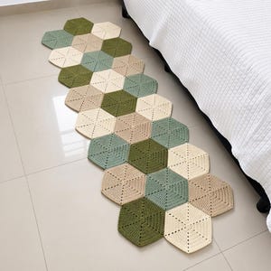 Puede incluir: Una alfombra de pasillo larga y estrecha compuesta por múltiples motivos hexagonales de ganchillo unidos entre sí. La alfombra presenta una paleta de colores repetitivos de verde salvia, verde oliva, crema y beige, creando un patrón geométrico.