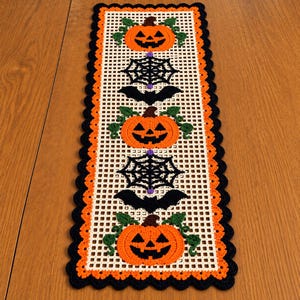 May include: A rectangular crochet table runner featuring a cream-coloured grid pattern with a scalloped border in orange and black. The design includes repeating motifs of orange jack-o'-lanterns with green vines, black spiderwebs, and black bats.