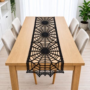 Pode incluir: Um caminho de mesa de crochê preto com um padrão repetitivo de teia de aranha. O caminho é retangular com uma borda festonada e está centralizado ao longo de uma mesa de jantar de madeira clara, exibindo um trabalho intrincado de fios.