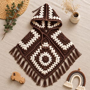 Op de afbeelding: Een poncho met capuchon gemaakt van donkerbruin en crèmekleurig garen in een granny square haakpatroon. Het kledingstuk heeft een puntige capuchon met bijpassende koordjes en een zoom afgewerkt met lange, donkerbruine franjes langs de onderranden.