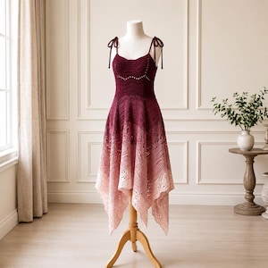 May include: A sleeveless crochet dress featuring a deep burgundy bodice that transitions into a lighter pink ombre skirt. The garment has thin shoulder straps with tie details, a fitted waist, and an asymmetrical handkerchief hemline.