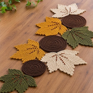 May include: A collection of crocheted table decor featuring several maple leaf shapes in cream, mustard yellow, and olive green, interspersed with circular dark brown coasters, all arranged in a scattered pattern on a wooden surface.