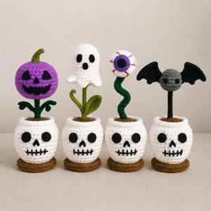 Könnte beinhalten: Vier gehäkelte Halloween-Dekorationen in weißen, totenkopfförmigen Töpfen. Die Figuren umfassen einen violetten Kürbis, ein weißes Gespenst, einen Augapfel an einem grünen Stiel und eine graue Fledermaus mit schwarzen Flügeln aus Garn.