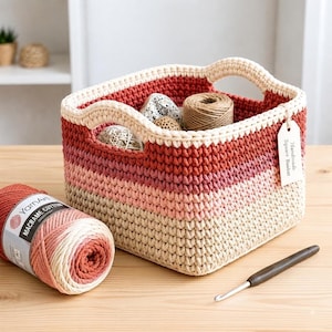 May include: A square crochet basket featuring a gradient of colors from dark red to light beige, with integrated handles and a white tag labeled Handmade Square Basket. A ball of matching cotton yarn and a crochet hook are placed beside the basket.