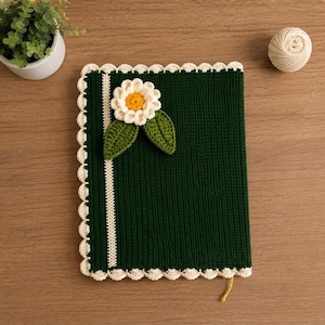 May include: A dark green rectangular crochet book cover featuring a white scalloped border, a vertical white stripe, and a three-dimensional white flower with a yellow center and two green leaves attached to the front surface of the cover.