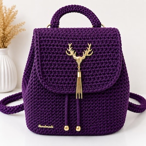 May include: A deep purple crochet backpack featuring a top handle and a flap closure. A gold-toned metal deer head emblem with a dangling chain tassel is centered on the flap. The bag includes a drawstring closure with gold-toned beads and a small tag.