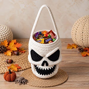Könnte beinhalten: Ein cremefarbener Häkelkorb in Form eines Totenkopfs mit einem einzelnen Henkel. Der Totenkopf hat schwarze gehäkelte Augenhöhlen, eine kleine dreieckige Nase und einen breiten grinsenden Mund mit Zähnen. Der Korb ist mit verschiedenen Riegeln gefüllt.