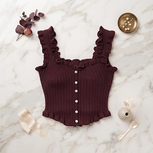 Peut inclure: Un débardeur en tricot bordeaux foncé doté d'une encolure carrée, de larges bretelles à volants et d'une patte de boutonnage avant avec six boutons blancs nacrés. Le vêtement présente un corsage ajusté à texture côtelée et un ourlet à volants.