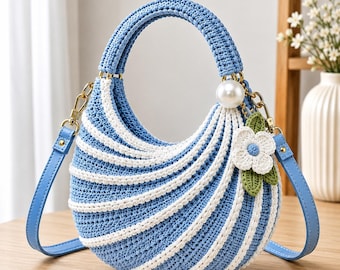 May include: A crescent-shaped handbag crafted from blue and white crocheted yarn in a striped pattern. The bag features a single top handle, a detachable blue shoulder strap with gold-tone hardware, a large white pearl bead, and a crocheted flower charm.