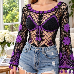 Op de afbeelding: Een zwart gehaakte top met lange mouwen, een wijde halslijn en uitlopende mouwen versierd met paarse bloemmotieven. Het kledingstuk heeft een zwart bralette-achtig lijfje met paarse rand en een striksluiting in het midden, gecombineerd met een lichtblauwe spijkerbroek.