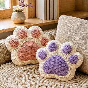 Può includere: Due cuscini all'uncinetto a forma di zampa di animale. Uno presenta una base color crema con un grande cuscinetto centrale rosa e quattro cuscinetti più piccoli. Il secondo ha una base color crema con un grande cuscinetto centrale viola e quattro cuscinetti più piccoli.