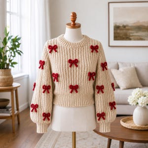 May include: A beige ribbed knit sweater featuring a crew neckline and long sleeves, decorated with multiple small, dark red yarn bows scattered across the front and sleeves. The garment is displayed on a mannequin torso against a neutral background.
