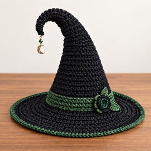 May include: A black crocheted pointed hat with a wide brim, featuring a dark green band around the base adorned with a crocheted flower and leaves. A gold-toned crescent moon charm with a small green bead hangs from the tip of the hat.