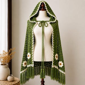 Pode incluir: Uma capa com capuz em crochê verde-floresta com padrão de malha aberta, borda festonada em tom creme e grandes motivos de margaridas brancas com centros amarelos. A peça inclui longos cordões trançados com borlas e uma bainha com franjas.