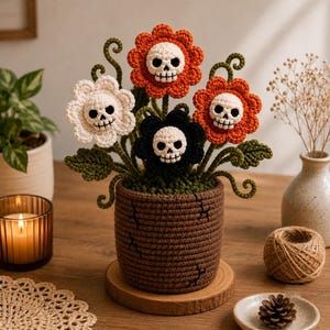 Könnte beinhalten: Eine gehäkelte Topfpflanze mit vier Blüten in Weiß, Orange und Schwarz, die jeweils in der Mitte einen weißen Totenkopf haben. Die Blumen haben grüne Stiele und Blätter und stehen in einem braunen, strukturierten Topf mit schwarzen Rissen.