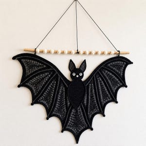 May include: A black and dark gray crocheted bat wall hanging suspended from a light wooden dowel. The dowel features a row of small, round, cream-colored crocheted beads. The bat has pointed ears, white circular eyes, and textured wings.