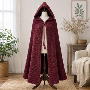 Op de afbeelding: Een vloerlange, diepbordeauxrode cape met capuchon, gemaakt met een getextureerd haakpatroon. Het kledingstuk is voorzien van een puntige capuchon, een enkele houten knoopsluiting bij de halslijn en twee lange, dunne gevlochten koorden met kwastjes.