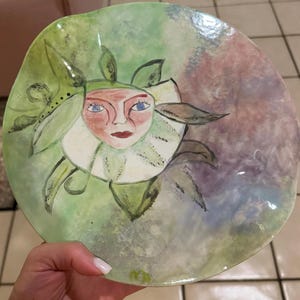 May include: A round ceramic plate with an irregular, wavy edge. The surface features a hand-painted illustration of a face surrounded by green leaves and petals. The background is a blend of soft green, purple, and blue watercolor-style washes.