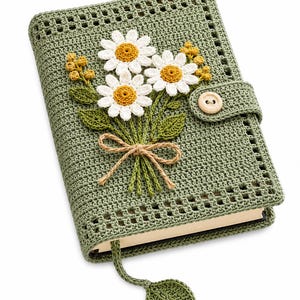 Puede incluir: Una funda de libro tejida a ganchillo en color verde salvia con un ramo de tres margaritas blancas de centro amarillo, pequeños capullos amarillos y hojas verdes. Un lazo de cuerda marrón ata los tallos. Un botón de madera asegura la funda y un marcapáginas en forma de hoja cuelga debajo.