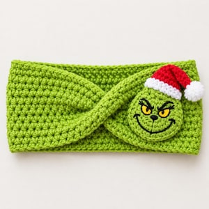 May include: A lime green crochet headband featuring a twisted front design. Attached to the right side is a circular green crochet patch depicting a character face with yellow eyes, a black stitched smile, and a red Santa hat with a white pom-pom.