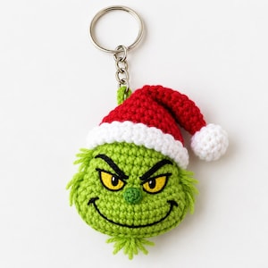 May include: A crochet keychain featuring a green character head with a mischievous expression, yellow eyes, and a wide black stitched smile. The character wears a red and white Santa hat with a white pom-pom, attached to a silver metal ring.