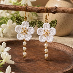 May include: A pair of dangle earrings featuring white crocheted flowers with green crocheted leaves, gold-tone metal centers, and a vertical drop of three small white faux pearls suspended from each flower on gold-tone metal earring hooks.