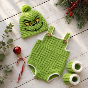 May include: A crochet set consisting of a bright green beanie with a pom-pom and a Grinch face, a green romper with white trim and two brown buttons on the straps, and a pair of matching green and white booties with a soft white cuff edge.