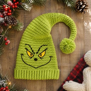 May include: A bright lime green crocheted slouchy hat featuring a long, curved tail with a matching pom-pom at the end. The front of the hat displays a black embroidered graphic of a mischievous face with sharp, yellow-colored eyes.