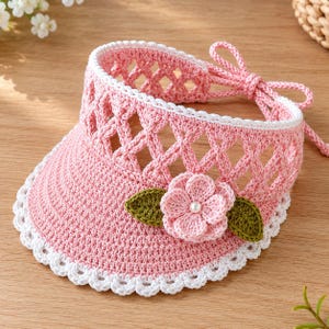 May include: A light pink crochet sun visor featuring a white scalloped trim along the brim and a lattice-patterned crown. The visor is decorated with a central pink crocheted flower with a small white pearl center and two green leaves.