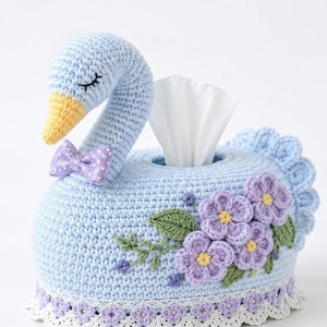 May include: A light blue crocheted tissue box cover shaped like a swan with a yellow beak, a purple polka dot bow, and purple crocheted flowers with green leaves on the side. The base features a white lace trim with small purple flower details.