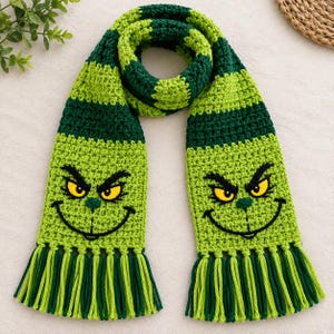 May include: A crocheted scarf featuring alternating horizontal stripes of dark green and lime green yarn. The ends of the scarf display a stitched face with yellow eyes, black eyebrows, and a wide black smile, finished with long green fringe.