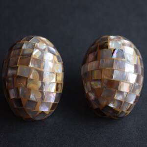 May include: Two oval-shaped decorative objects featuring a mosaic pattern of iridescent, multi-colored mother-of-pearl tiles. Each piece is constructed with small, square-cut shell segments arranged in a grid, displaying shades of brown, gray, and white with a reflective, pearlescent finish.
