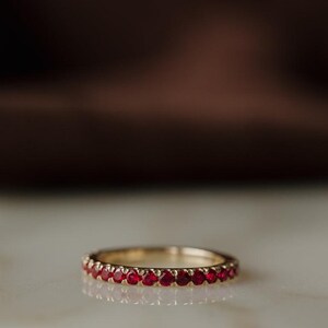 Könnte beinhalten: Ein goldener Ring mit einer durchgehenden Reihe kleiner, runder, tiefroter Rubine in einer gemeinsamen Krappenfassung. Der Ring hat eine polierte metallische Oberfläche und ein schmales Profil, das eine gleichmäßige Anordnung der Steine zeigt.