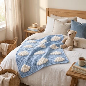 May include: A light blue crocheted blanket featuring raised white bobble clouds across the surface. The blanket is draped over a bed next to a beige knitted teddy bear. The texture of the yarn is visible throughout the square-shaped design.