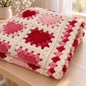 Può includere: Una coperta all'uncinetto piegata caratterizzata da un motivo quadrato ripetuto con cuori centrali in tonalità di rosso e rosa, bordati da filato color crema. Il tessuto presenta una costruzione testurizzata fatta a mano con cuori alternati.