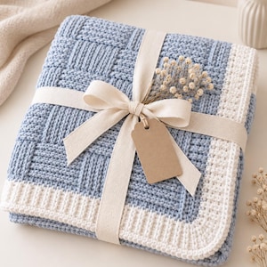 May include: A folded light blue and white knit blanket featuring a basketweave texture and a ribbed border. The blanket is tied with a cream-colored fabric ribbon, adorned with a small sprig of dried white flowers and a blank brown paper tag.