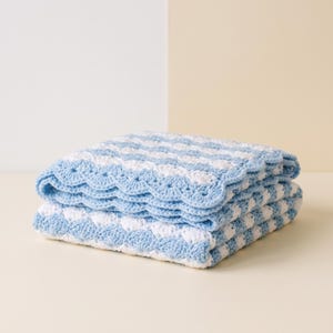 May include: A folded crochet blanket featuring alternating horizontal stripes of light blue and white yarn. The blanket is crafted with a textured shell stitch pattern and finished with a decorative scalloped edge in light blue yarn.