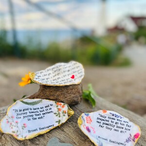 May include: Three oyster shells with gold-painted edges, each featuring printed text and floral illustrations. One shell displays a red heart, while the others contain quotes by Ernest Hemingway about journeys and distance, set against a natural background.