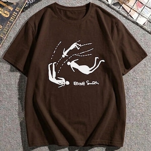 May include: A brown short-sleeved t-shirt featuring a white graphic print of three stylized figures in motion with dashed lines indicating trajectory. The text Elliott Smith is printed in white below the figures on the front of the garment.