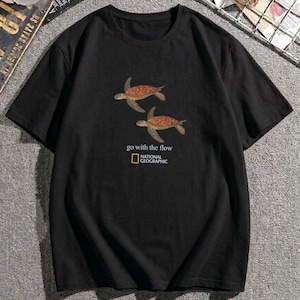 Puede incluir: Una camiseta negra de manga corta con un gráfico de dos tortugas marinas nadando en tonos marrones y naranjas. Debajo de las tortugas, el texto dice go with the flow, seguido del logotipo de National Geographic en su rectángulo amarillo.