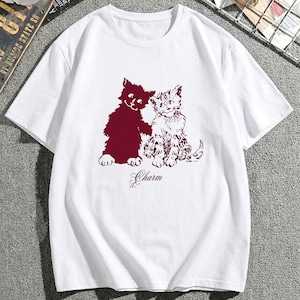 May include: A white short-sleeved t-shirt featuring a graphic print of two cats in a dark maroon colour. One cat is a solid silhouette, while the other has detailed fur markings. The word Charm is printed in a cursive script font below the cats.