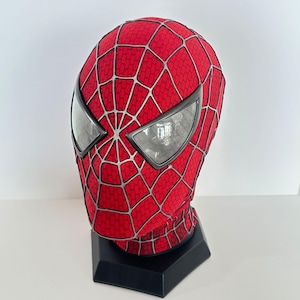 May include: A red full-head mask featuring a raised silver web pattern across the entire surface. The mask includes two large, angular, reflective silver-rimmed eye lenses. It is mounted on a black hexagonal display stand for structural support.