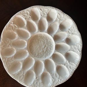 May include: A white ceramic egg serving plate featuring a circular central area surrounded by eighteen individual oval-shaped indentations. The surface of the plate is decorated with a textured leaf and vine pattern throughout the rim.