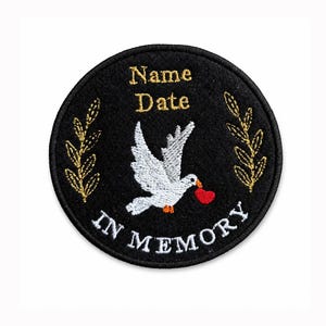 May include: A circular black embroidered patch featuring a white dove carrying a red heart in its beak. The text "Name" and "Date" appear in gold above the dove, while the words "IN MEMORY" are stitched in white along the bottom edge.