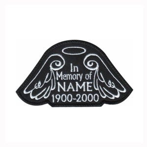 May include: A black embroidered patch shaped like a pair of angel wings with a halo above. The white stitching features the text In Memory of NAME and the dates 1900-2000 centred between the wings. The patch has a clean, arched top edge.