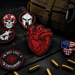May include: A collection of tactical morale patches including a red anatomical heart, a skull in a red helmet, a spartan helmet, a compass rose with the text I'M NOT LOST I'M EXPLORING, and a skull with an American flag pattern, arranged on a dark surface.