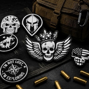 May include: A collection of black and white morale patches featuring various designs including a skull with wings and a crown, a Spartan helmet, a compass rose with the text I'M NOT LOST I'M EXPLORING, and a skull with an American flag pattern.